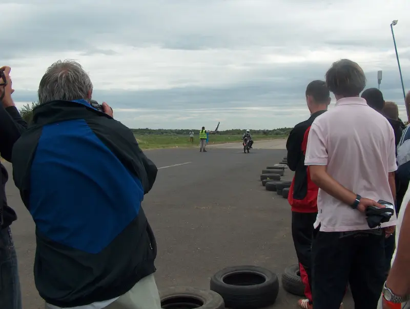 The motorcycles arrive at the finish line one after the other on the race track. People are waiting on both sides to take pictures of them.