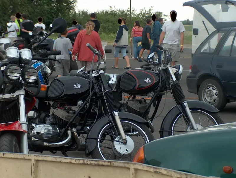 Kunmadarasi 24 órás motorverseny helyszíne. 2 db fekete Pannonia motorkerékpár van középen. Mögötti számos, színes ruhába öltözött emberek. Jobb oldalon egy kék autó kinyitott csomagtér ajtóval.