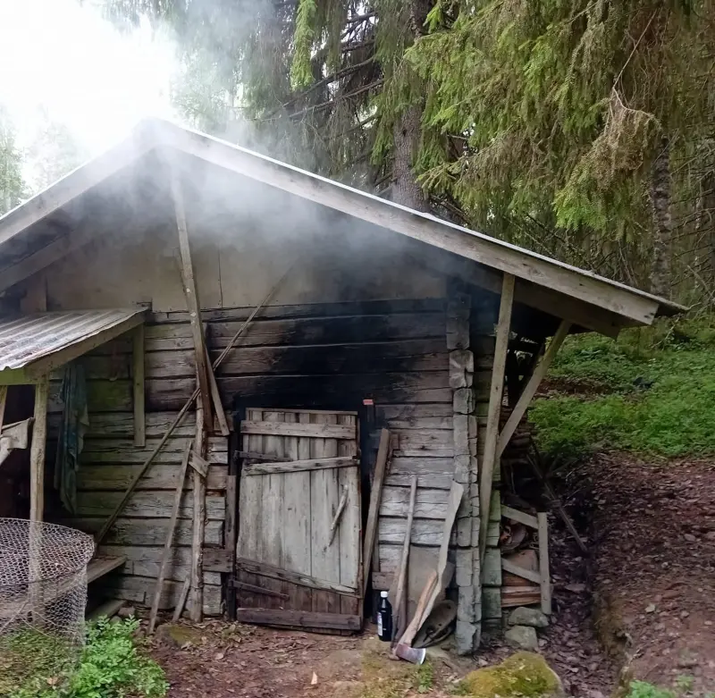 Rear part of an externally heated sauna The rear part of the sauna is visible. This is where the heating takes place. Everything is made of wood. There are some tools next to the wall, e.g., an axe. In the upper right corner, pine branches hang down toward the roof.