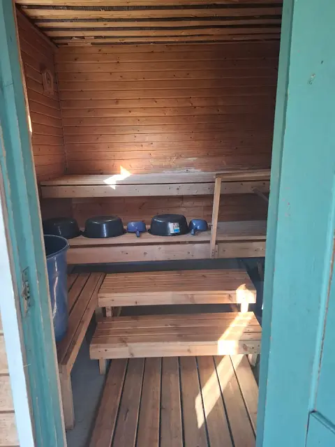 Egy szauna belső tere. A képen fa burkolat mindenhol.Fa a oadló, a mennyezet, az ülő- és fekvőrész. CSupán az ajtó keret égszínkék. Két lépcső van középen, mely a fekvő részig visz. Ez alatt polc, rajta több sötétkék tároló és merítő eszköz. Balra az ajtónál egy részben látszódó ülő rész.