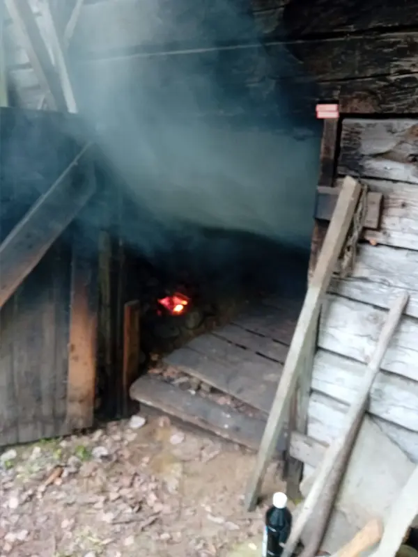 Külső fűtésű szauna A szauna nyitott hátsó ajtaja látszik. Itt zajlik a fűtés. Balra egy kis tűz, felül füst és kormos tetőrész. Minden fából van.