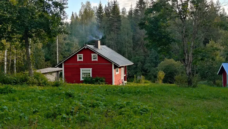 In the forest, on a green clearing, there is a red cottage with two small and one larger white window. There are trees on the left and in the background. Smoke is coming out of the chimney. A very low, gray structure can also be seen in front of the cottage, perhaps an outdoor storage shed. On the right, in the middle, a small piece of the blue roof of a building hangs into the picture. The sky is blue with few clouds.