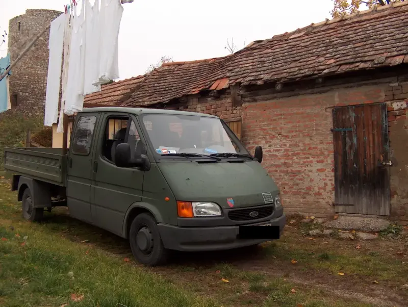 Siklós-a vásárlás helyszíne A siklósi vár tövében, egy nagyon hosszú, hullámos tetejű, rozoga épület. Előtte egy katonai zöld színű kis teherautó. ruhák kiterítve az udvaron.