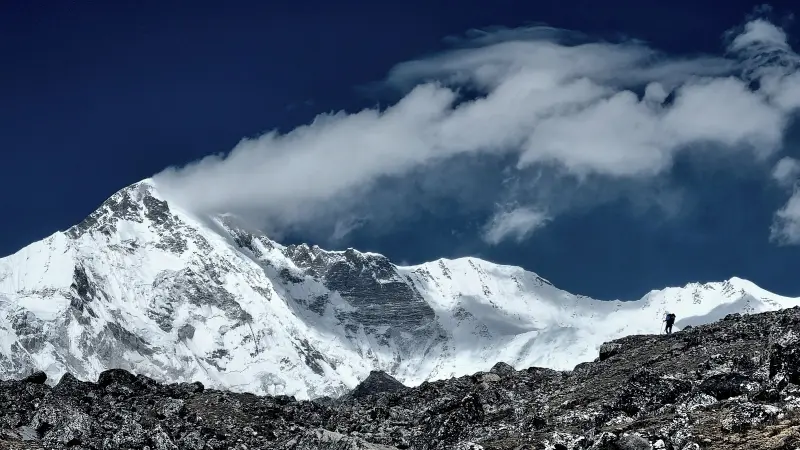 In the foreground of the picture is grayish, rising ground. On the right, at the top, in the distance, is the barely visible figure of a person. In the background is a white mountain peak.