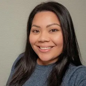 A young woman with black, long hair, dark skin, and a smile, wearing a blue long-sleeved sweater.