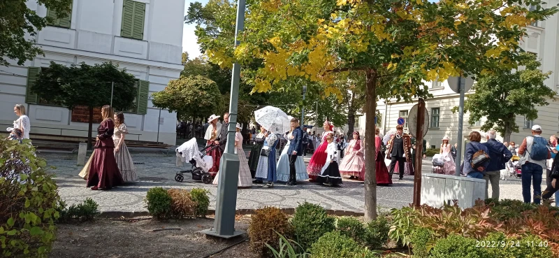 People dressed in elegant old-fashioned long dresses parade down the street. One carries a large white parasol, another pushes an old-fashioned pram. Others watch them from the right side of the street.