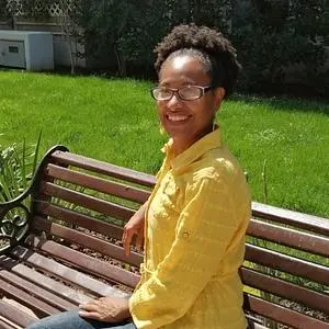 A woman with brown skin, very curly hair, and glasses. She is wearing a yellow blouse. She is sitting on a wooden bench outdoors, turned slightly to the side.