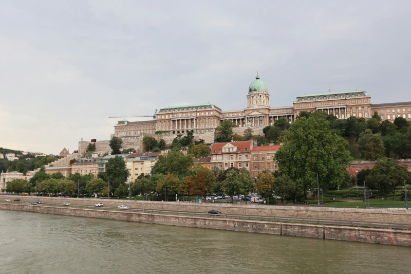 Duna, háttérben a rakpart, házakkal, fákkal. Felette emelkedik a Budai Vár.  Majdnem a teljes képen végig húzódik, közepén kupola.