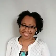 A smiling woman with brown skin, curly hair, and glasses. She is wearing a white cardigan. Around her neck is a necklace with a large blue pendant.