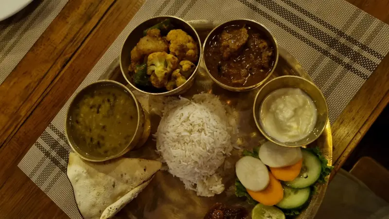 A small brown tablecloth with a brown checkered tablecloth on top. On a round, golden tray, there are four small round bowls containing different foods. In the middle, rice is piled up in a cone shape. On the right side of the tray, there are cucumbers, and on the left side, there is triangular bread.