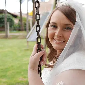 A smiling young woman sitting on a swing outdoors. She is wearing a transparent, light-colored veil-like scarf on her head.