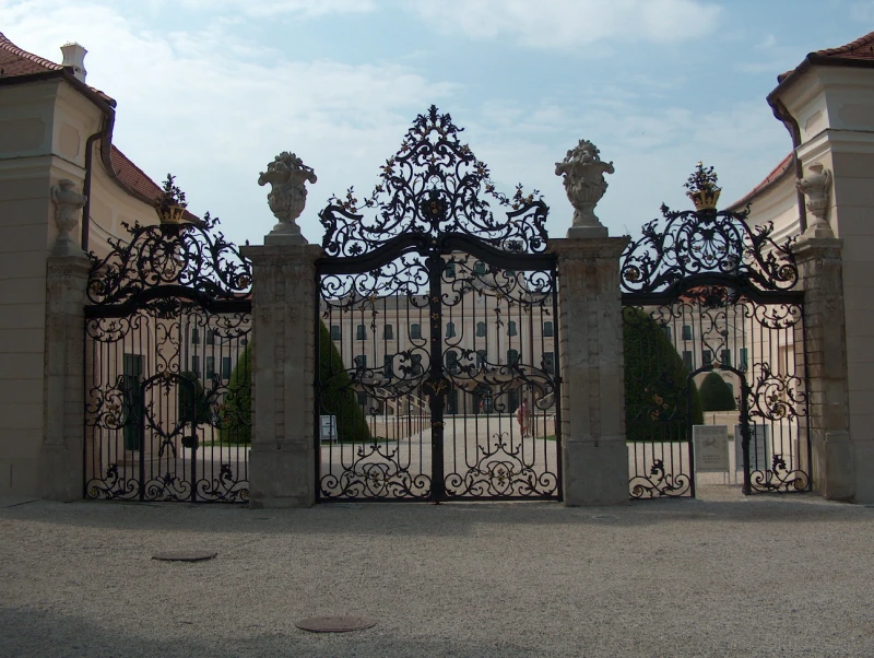 The ornate metal gate of Fertőd Castle, behind which the castle can be seen. The gate has three pairs of wings, with the two central wings slightly larger than the others. Each section of the gate is topped with a metal, branch-like decoration that rises high in the center to form a dome. There are stone columns on either side of the central gate section. On top of these are stone decorations resembling hourglasses, complemented by decorations similar to those on the upper metal section of the gate. One of the wings of the right-hand gate is open.