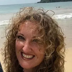 A young woman with a smile and light, curly hair. She is outdoors, perhaps on a beach. Behind her, waves can be seen breaking on the shore.