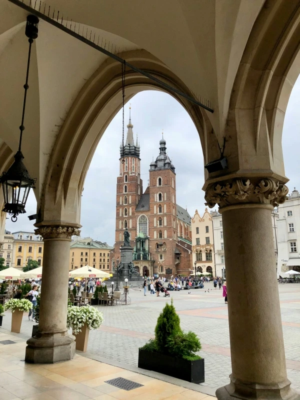 A picture of Krakow's main square. In the foreground is an ornate arcade, through which a brown church with two towers can be seen. Next to it, on the right and left, are multi-story houses. There are people in the square. In several places there are pavilions crowded with people.