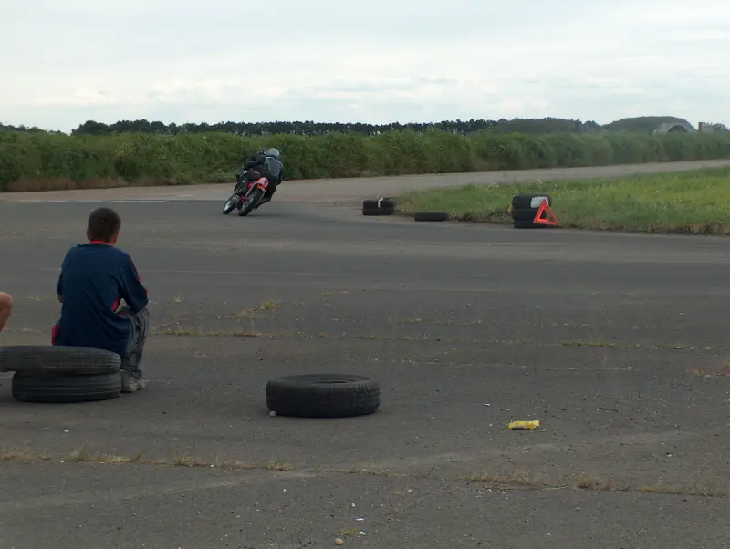 Veterán motorverseny .Balra a távolban három motoros halad a kanyar felé. Előtérben zöld terep. Távolban jobbra fák. Bal oldalon domb látható