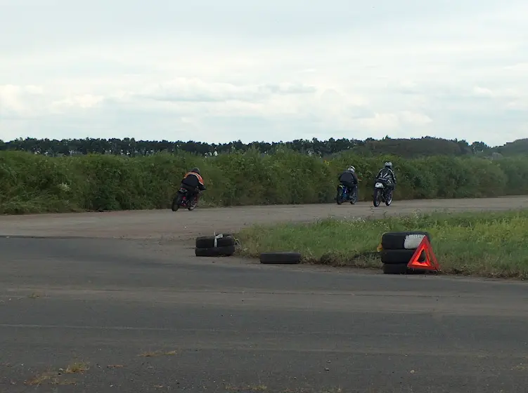 Veterán motorverseny .Balra a távolban három motoros halad a kanyar felé. Előtérben zöld terep. Távolban jobbra fák. Bal oldalon domb látható