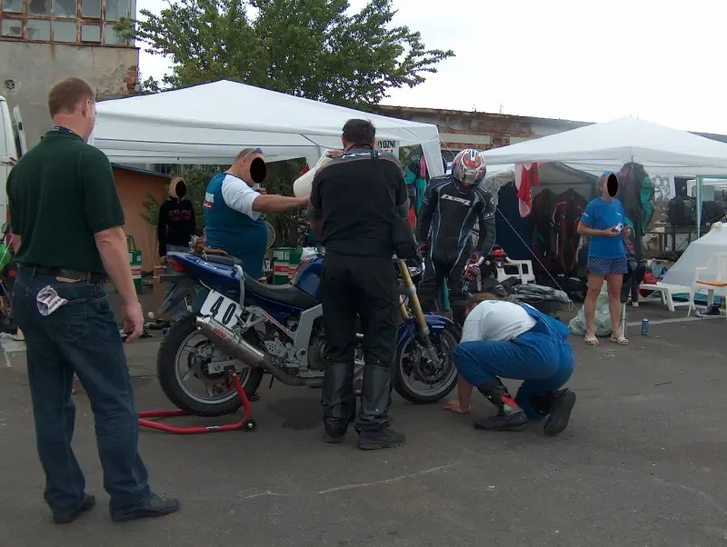 A pályán kívül, a sátrak előtt többen próbálnak megszerelni egy motort. Egy másik motoros is nézi a műveletet.