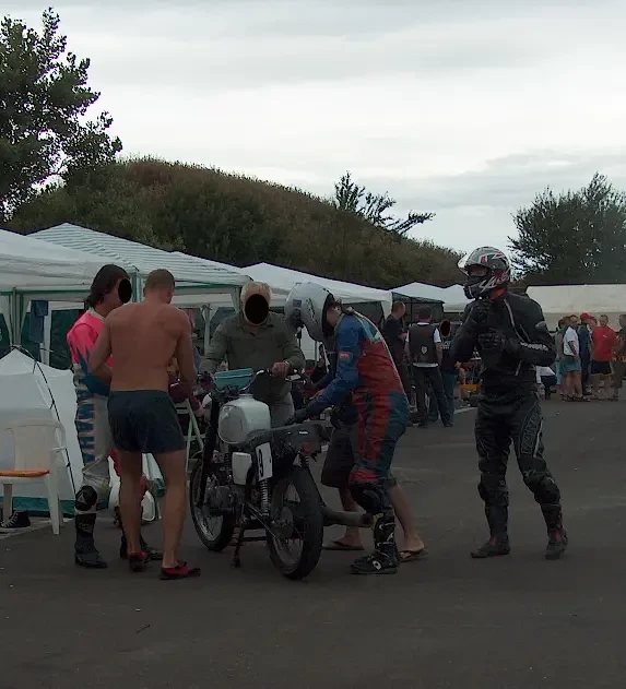A pályán kívül, a sátrak előtt többen próbálnak megszerelni egy motort. Egy másik motoros is nézi a műveletet.