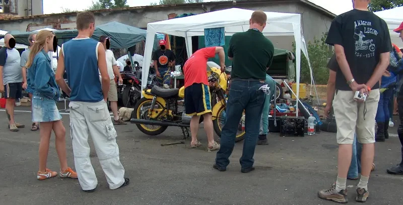 A pályán kívül, egy sátor előtt többen próbálnak megszerelni egy sárga motort. Körülöttük több ember , akik nézik a műveletet.