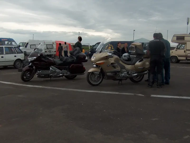 Két nagy motorkerékpár. Egyik fekete, a másik arany színű. Mellette emberek, mögöttük autók.