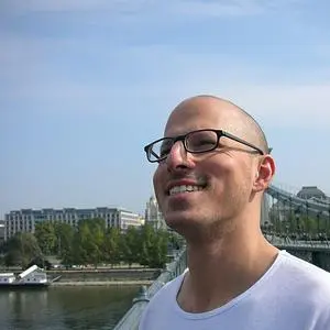 Fehér polót viselő, szemüveges, kopaszodó fiatal férfi. Jobb oldali profilját mutatja. Háta mögött egy folyó látszik. Jobb oldalon egy híd, balra a folyó mögött, a távolban többszintes emeletes házak.