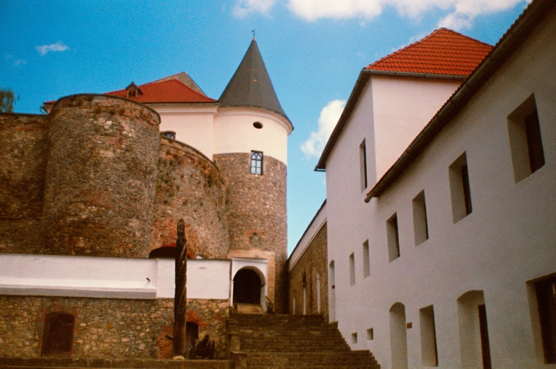 On the left side of the picture is an ancient, multi-circular part of the castle. On top of one of them is a small tower with a green dome. On the right side is a light-colored two-story old building with a third level in the middle, topped with a tent roof.