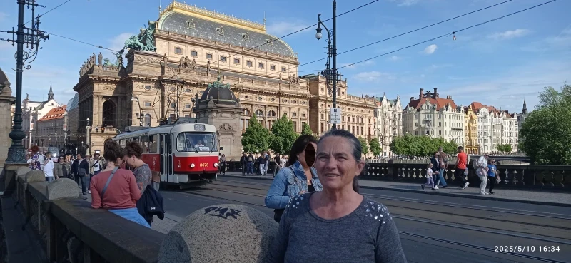 Egy forgalmas út, előterében állok. Mögöttem balra egy piros villamos érkezik az úton. Háttérben többszintes díszes épület. Tetején szobrok, és egy dísz lovas fogat.
