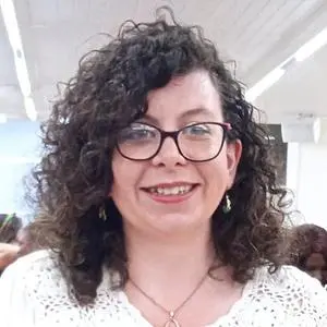 A smiling woman with brown, curly hair and glasses. She is wearing a white lace crocheted blouse.