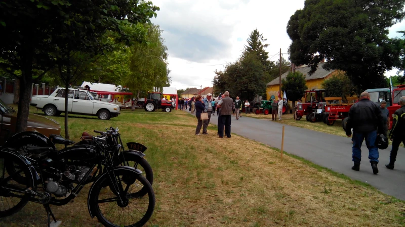Motoros találkozó helyszíne. Bal oldalon lent több régi motor, távolabb egy fehér régi autó. Az utcán mindenhol emberek jönnek-mennek. A képen bal oldalon a háttérbe és jobb oldalon 2 traktor látható.