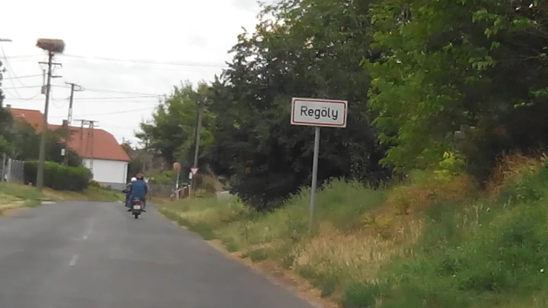 The boundary of the village of Regöly is marked by a sign. Two motorcyclists can be seen in the distance, riding away. There is a stork's nest on top of one of the electricity poles.