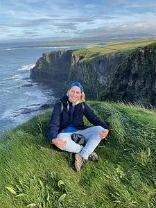 A woman sitting on the ground somewhere on a high seaside cliff. She crosses her legs at the ankles and holds both knees. She is wearing light blue jeans and a dark jacket. She has a blue scarf on. Behind her, you can see the sea and tall green cliffs jutting out from the shore into the sea.