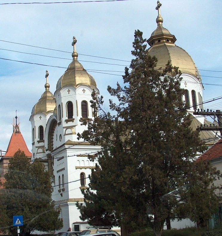 Orthodox church in Nagyszalonta, Romania. Three towers, white building with golden domes. Small arched windows all around the upper floor of each tower. Crosses on top of the domes.there is a huge arched window. The building has three decorative triangular structures on top, and in the middle, there is a church tower-like section. On the right side, there is a two-story building with a red exterior.