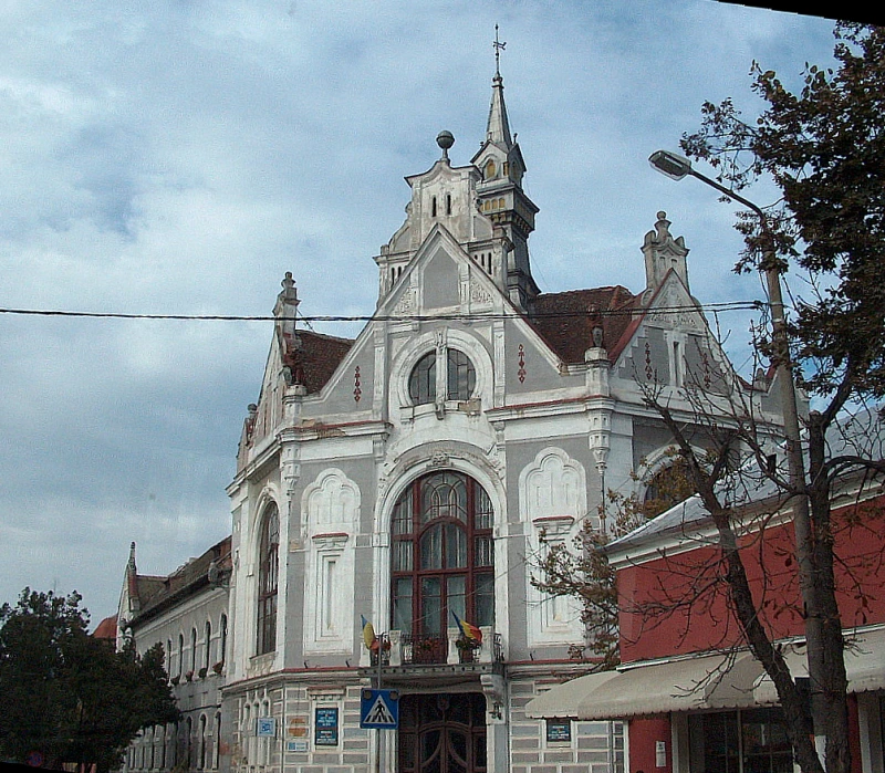 I took this photo of the Town Hall in Nagyszalonta during my travels abroad in Romania. It shows a two-story corner building with walls in a combination of grayish blue and white. On the upper floor, 