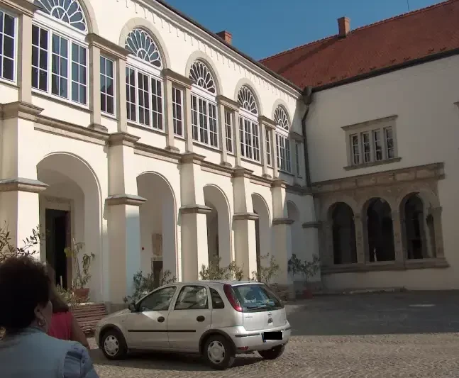 A képen bézs színű alul árkádos, felül ívelt tetejű, sok üveges ablakok. Az épület előtt egy kis, ezüst színű autó parkol.
