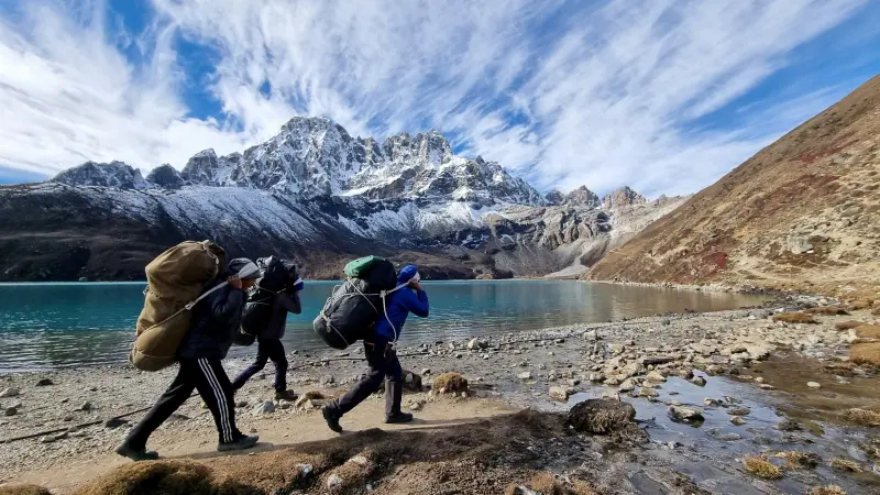 Three people are walking along the lake shore, carrying heavy backpacks. On the other side of the lake are very high white mountains.