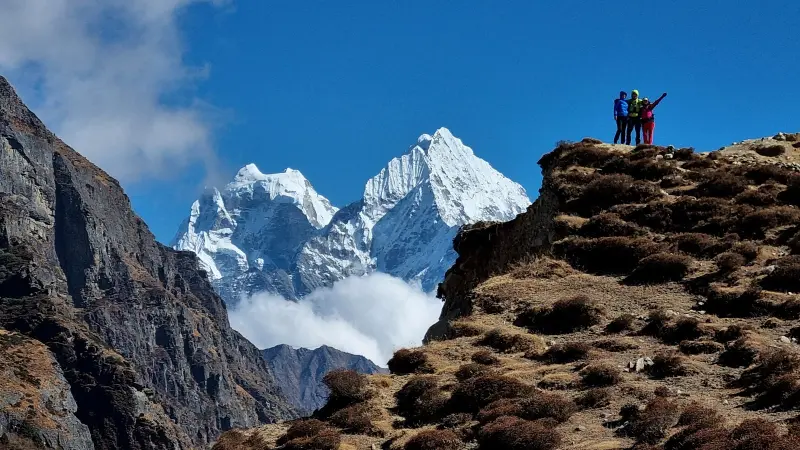Brown mountainsides are visible on both sides of the picture. A few people are cheering at the top of one of them. In the middle, there are white mountain peaks in the distance.
