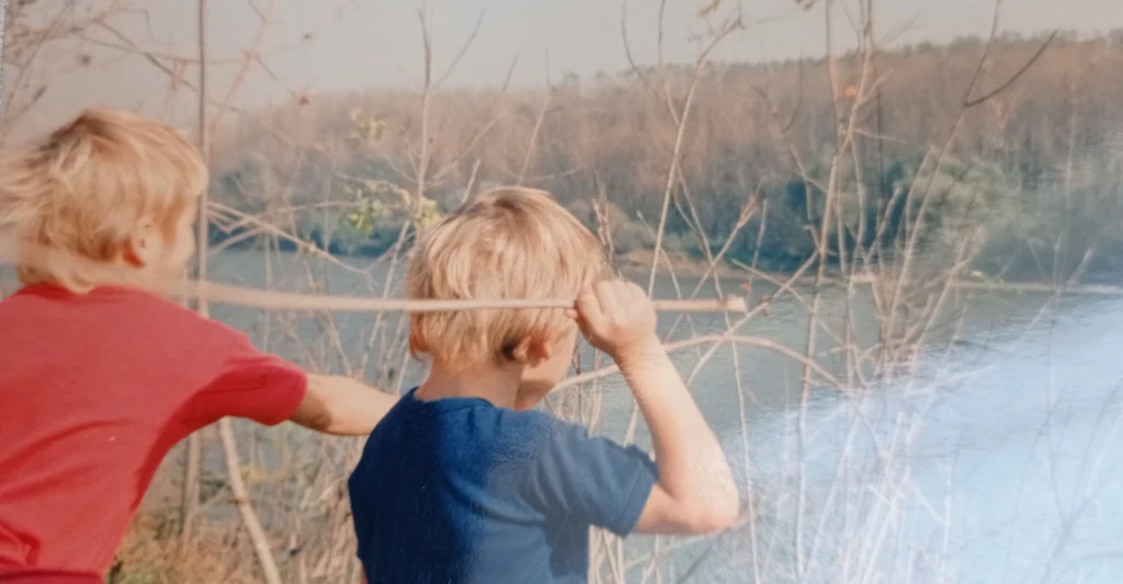 Tisza partján háttal ülve kb. 8 éves iker gyermekeim nád szálakkal próbálják eltalálni a vizet. Egyiken piros, a másikon kék póló van.Szemben látszik a túlsó part. 