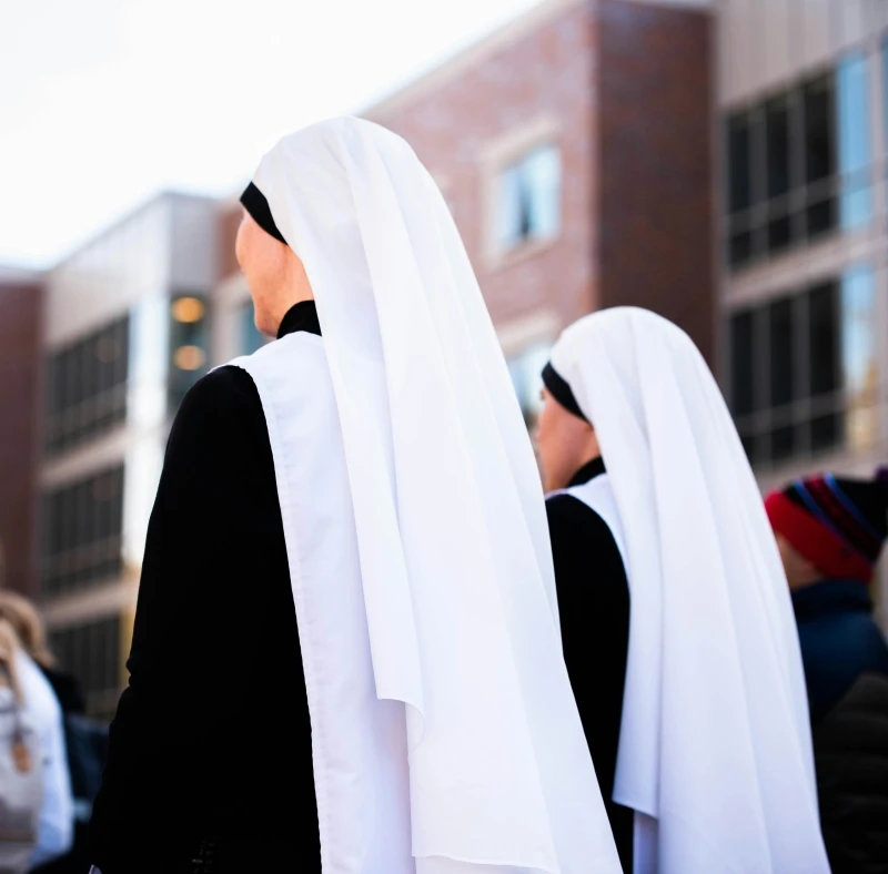 Two nuns can be seen from behind on the street. They are wearing blue dresses and white headdresses (bonnets).
