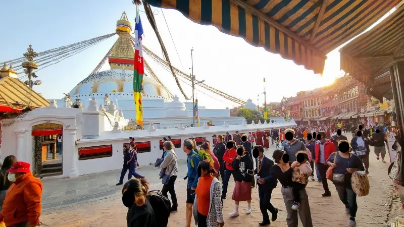 Emberek sietnek egy széles úton egy irányba. Háttérben talán egy hajó vagy egy díszes épület van. Ennek közepén egy hatalmas csúcsos arany színű felépítmény, körben piros dísszel. Középső rész csúcsából minden irányba kötelek. A kép jobb felső részén zöld-sárga csíkos ablak árnyékolók. Háttérben jobb oldalon húzódó emeletes házsor.
