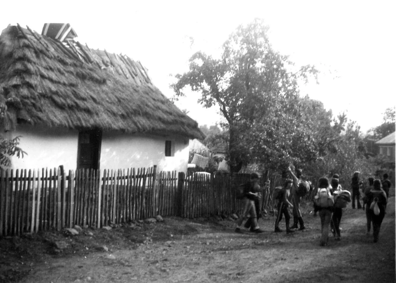 A group of young people are wandering through an old village. Only their backs are visible. On the left is a very old house with a thatched roof.