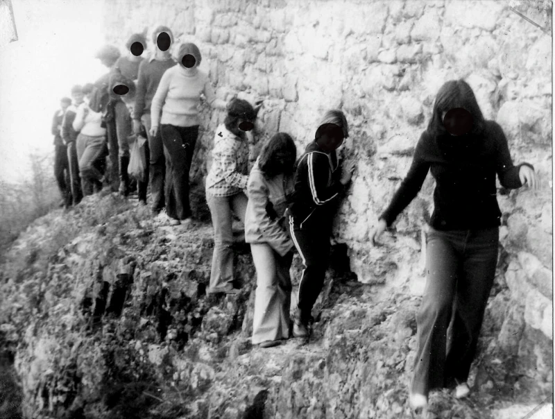 Young people walk one after another along the narrow path along the castle wall. The castle wall rises high beside them. They try to cling to the castle wall.
