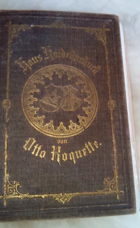 A brown old book with a gold-plated circular decoration on the upper part. Below this is a gold-plated semicircular inscription in Gothic letters: Otto Roquette.