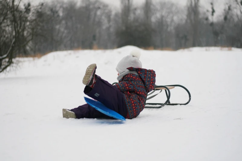 Winter landscape. A small child has just fallen off the sled. He is sitting in the snow next to it, one leg raised toward the sky.