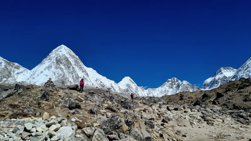 In the first part of the picture, there is a brown mountain peak. Several people are standing on it, far apart from each other. In the distance, there are very high white mountain peaks.