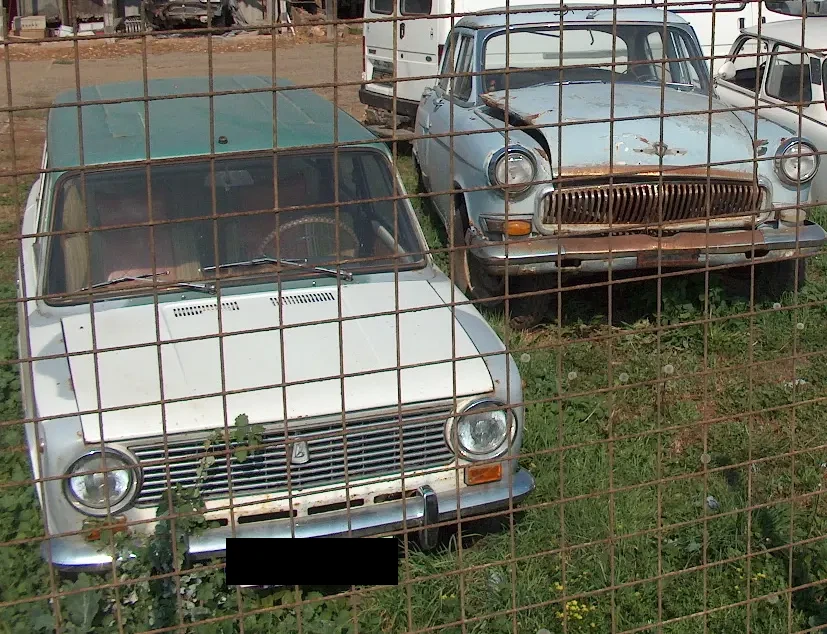Old white Lada and blue Volga cars behind a fence in Sárospatak.”