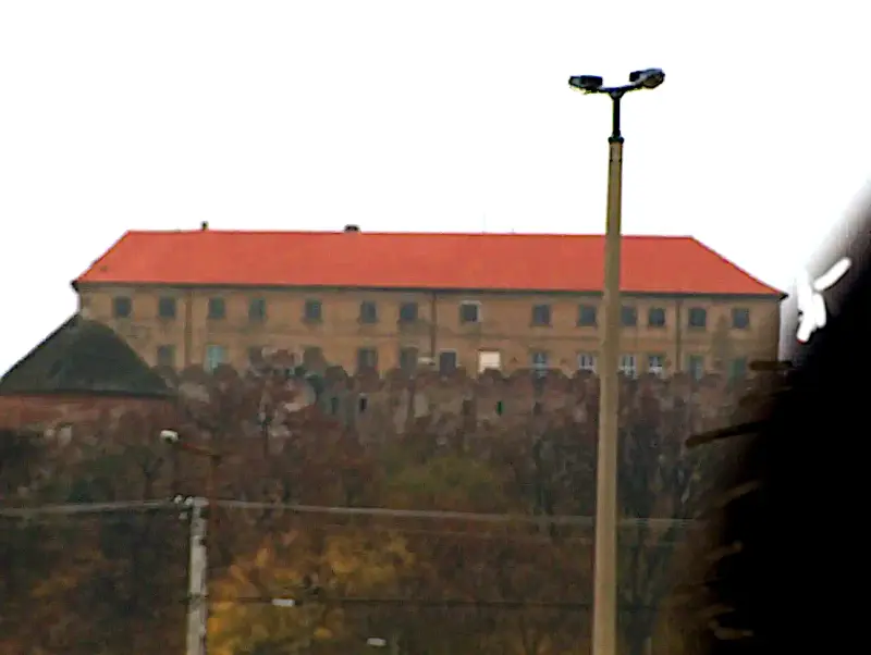 Siklós-A Vár a távolból A vár fő épülete látszik a távolban, teljes hosszában. Piros vártető, sárgás-barna falak, nagyon sok ablakkal. Előtte a régi várfalak. Előtérben egy új fajta utcai lámpa.